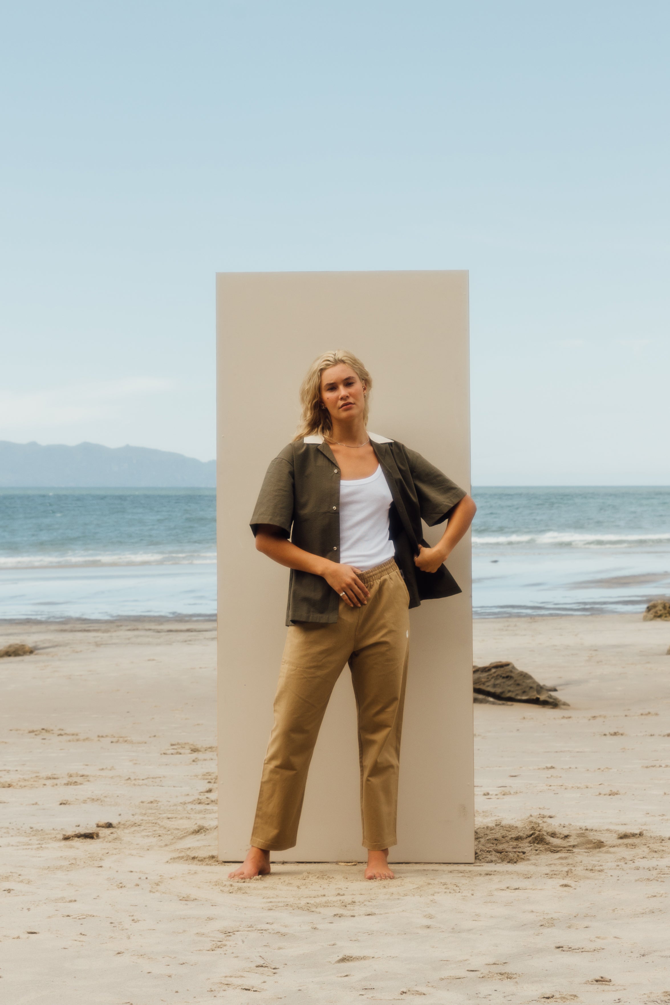 girl standing on beach in front of beige board wearing khaki shirt, white singlet, and beige pants