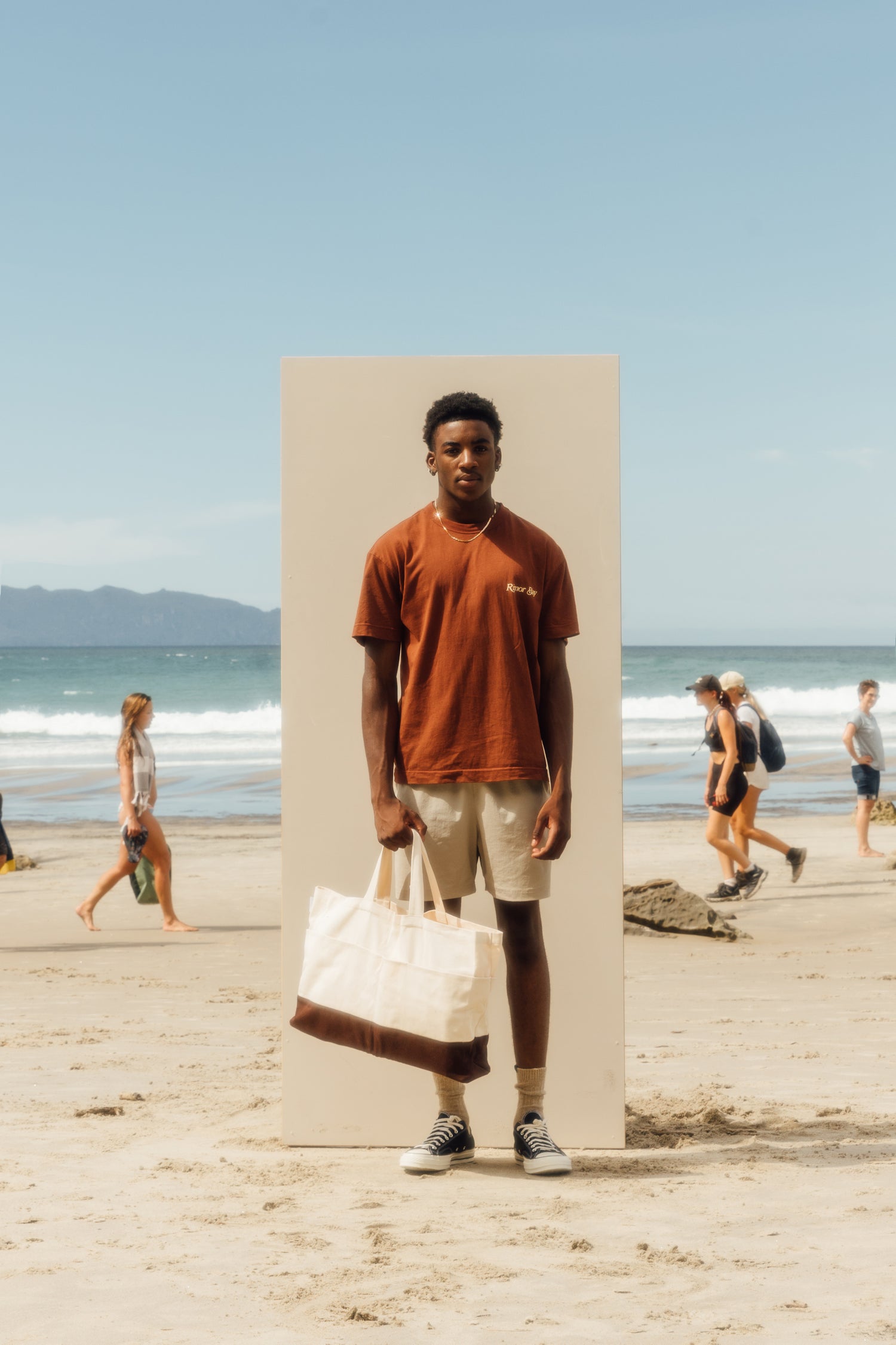 man standing on beach in front of beige board, wearing brown tshirt, gold chain, chucks, grey shorts, holding a beige bag 