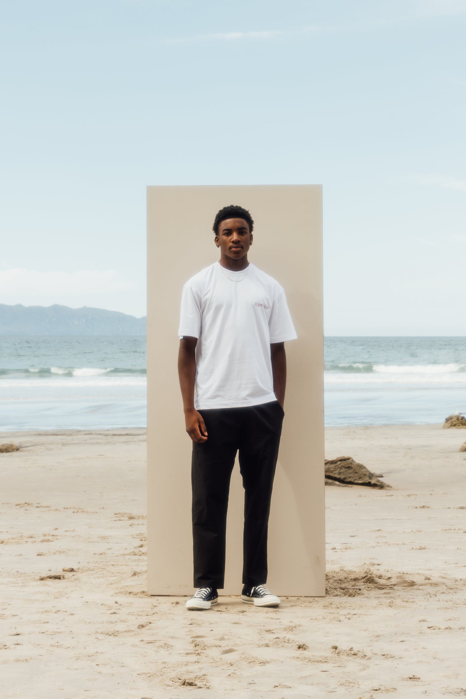 man standing on the beach in front of a beige board wearing white tshirt, black pants, and chucks