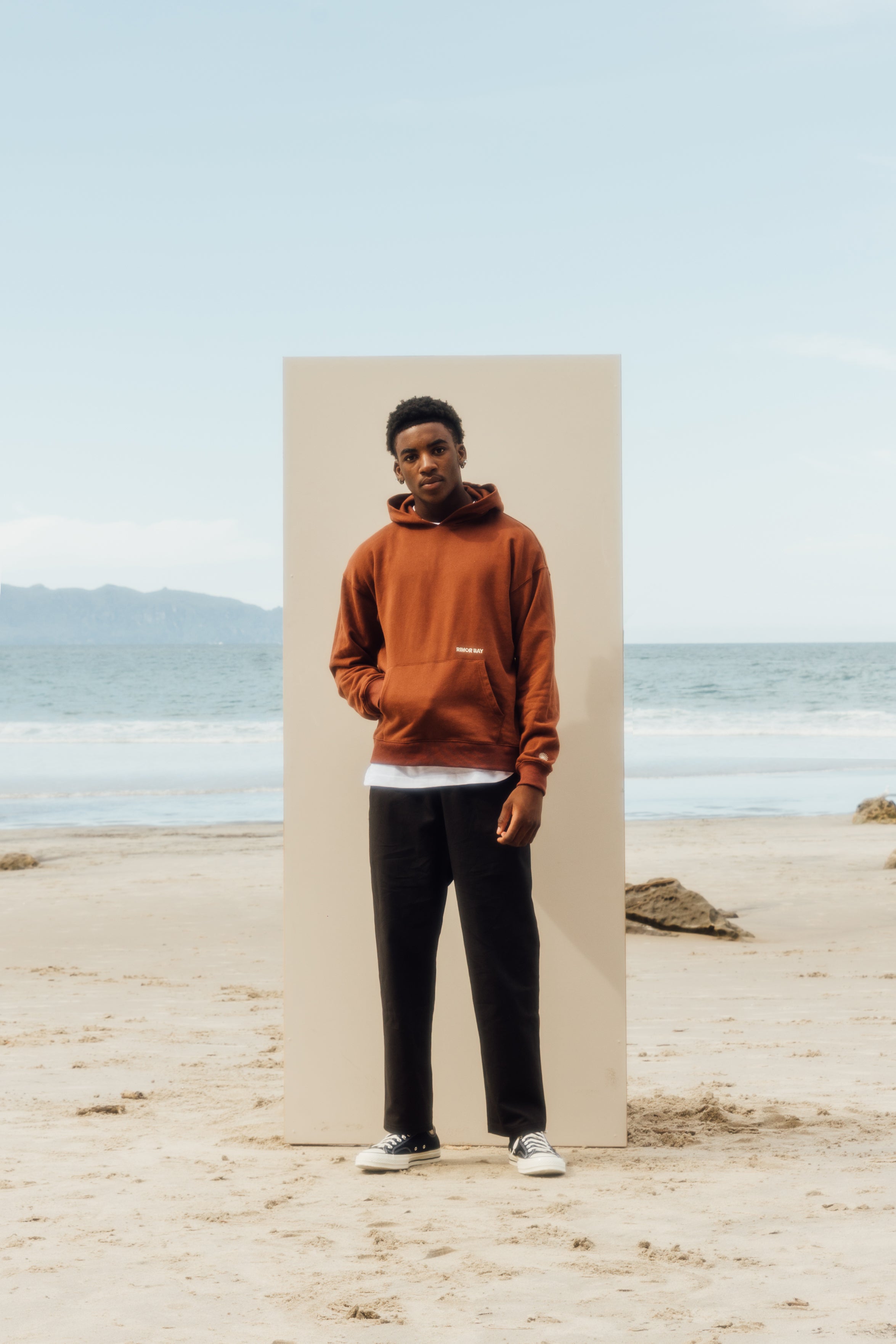 man standing on beach in front of a beige board, wearing brown hoodie white tshirt, black pants, and chucks