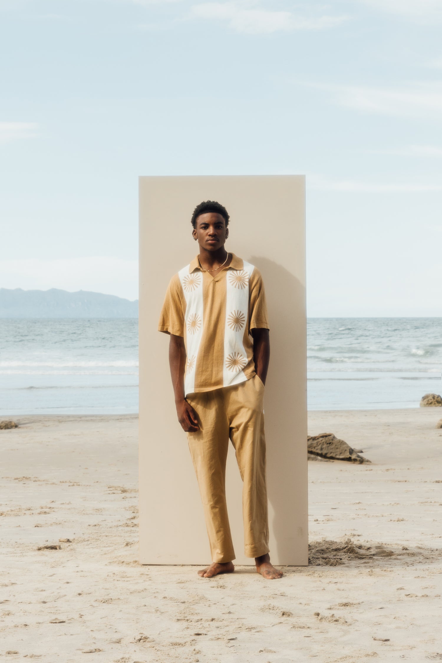man standing in front of beige board wearing knit top, and beige pants
